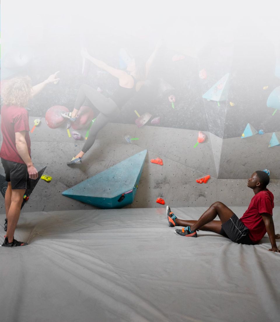 personas practicando escalada en un rocódromo con colchoneta gris acolchada que cubre el suelo para amortiguar caídas y entrenar con seguridad
