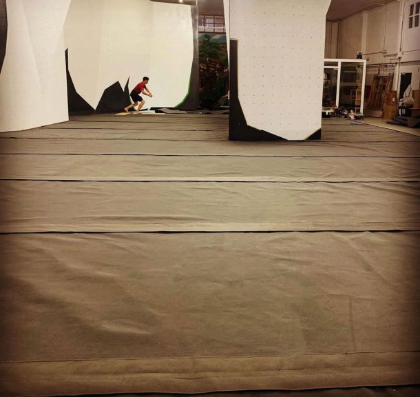 vista interior de un rocódromo con colchonetas grises cubriendo el suelo y una persona preparando una escalada
